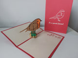 A 3D papercraft robin bird standing on a small green branch inside an open cream-colored greeting card with red borders. A red backing card standing behind it features a white bird illustration and the text 'to a great friend'.