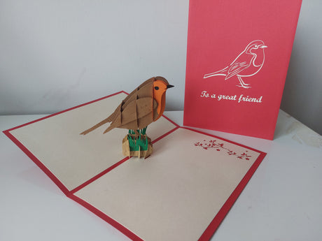 A 3D papercraft robin bird standing on a small green branch inside an open cream-colored greeting card with red borders. A red backing card standing behind it features a white bird illustration and the text 'to a great friend'.