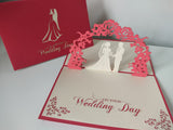 Wedding card with a 3D cutout of a bride and groom under an arch, on a red and white background.
